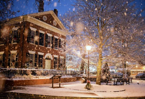 A historic brick building surrounded by snow, with a tree and street lamp illuminated in a winter evening scene.