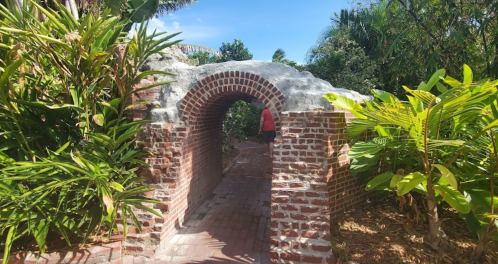 A brick archway surrounded by lush greenery, leading to a pathway in a tropical garden.