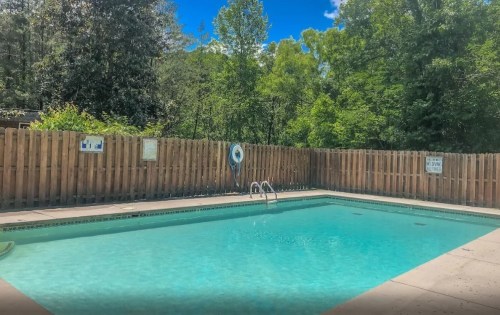 A clear swimming pool surrounded by a wooden fence and lush green trees under a bright blue sky.