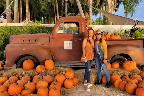 Two women pose beside a vintage truck surrounded by pumpkins in a sunny outdoor setting.