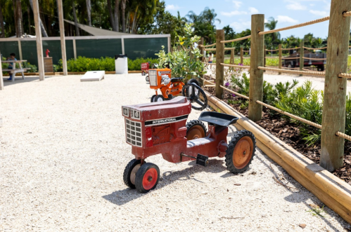 A small red toy tractor parked on gravel, with a second orange tractor visible in the background among greenery.