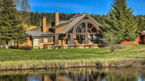 A large wooden house with a sloped roof, surrounded by trees, reflecting in a calm lake under a clear blue sky.