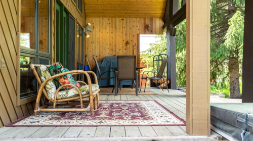 A cozy porch with a rattan chair, a patterned rug, and a dining set surrounded by wooden walls and greenery.