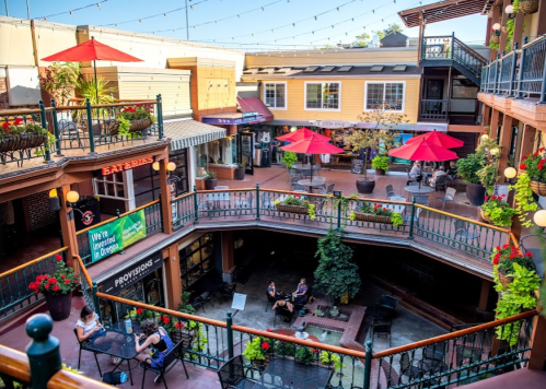 A vibrant courtyard with red umbrellas, greenery, and people enjoying a sunny day in a charming shopping area.