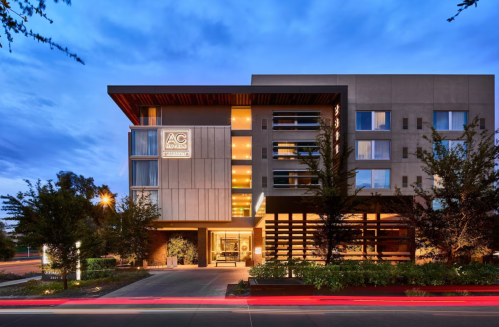 Modern hotel exterior at dusk, featuring large windows, greenery, and a welcoming entrance.