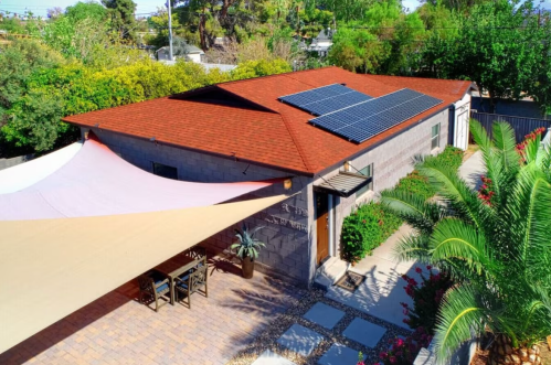 Aerial view of a house with a red roof, solar panels, and a shaded patio surrounded by greenery and landscaping.