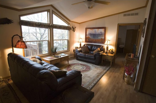 Cozy living room with leather sofas, a coffee table, and large windows overlooking a wooded area. Warm lighting and decor.