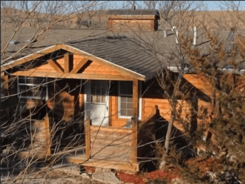 A wooden cabin with a porch, surrounded by trees and a clear sky in the background.