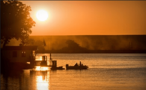 Sunset over a calm lake, with silhouettes of trees and a small boat near a dock. Warm orange and yellow hues in the sky.