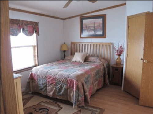 Cozy bedroom with a wooden bed, patterned quilt, lamp, and artwork on the wall, featuring natural light from a window.