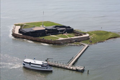 Aerial view of a historic fort on an island, with a boat approaching the dock and water surrounding the land.