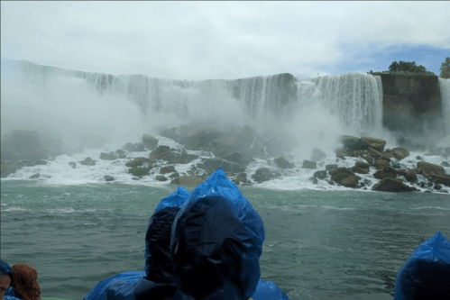 Visitors in blue ponchos stand near a powerful waterfall, with mist rising and rocks visible in the foreground.