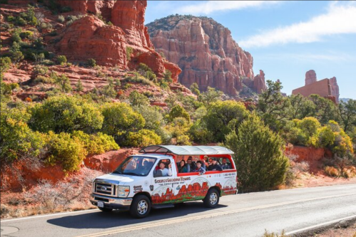 A tour van drives along a scenic road surrounded by red rock formations and greenery.