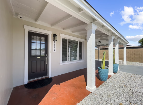 Front porch of a house with a dark door, large window, and potted plants, featuring a gravel pathway and blue sky.