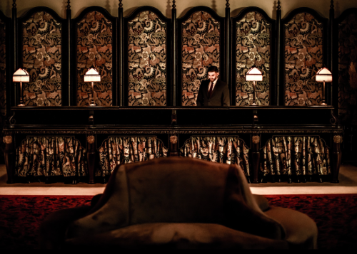 A dimly lit hotel reception with a man in a suit behind the counter, ornate decor, and a plush chair in the foreground.