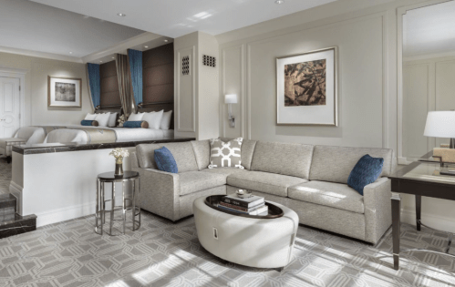A stylish hotel suite featuring a gray sectional sofa, a round coffee table, and a bedroom area in the background.