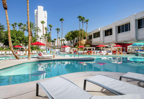 A sunny pool area with lounge chairs, palm trees, and red umbrellas, surrounded by a modern building.