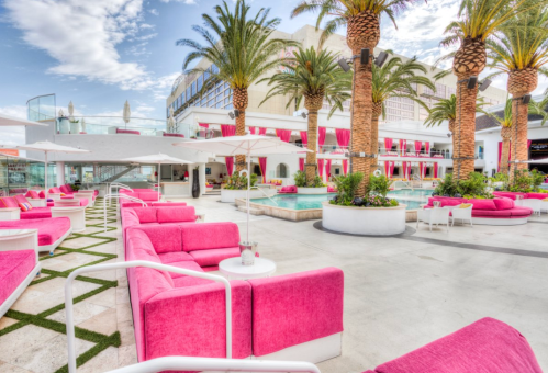 Vibrant outdoor lounge area with pink seating, palm trees, and a pool, set against a bright blue sky.
