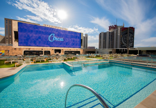 A sunny pool area with lounge chairs and a large screen displaying "Circa," surrounded by modern buildings.