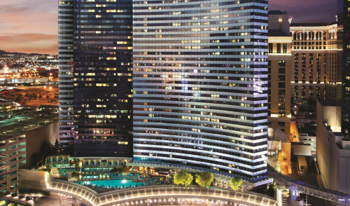 Aerial view of modern skyscrapers at night, with city lights and a pool area illuminated below.