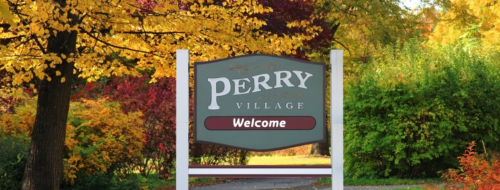 A welcome sign for Perry Village surrounded by vibrant autumn foliage.