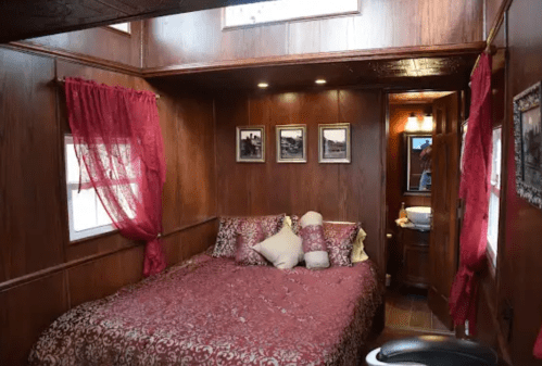 Cozy wooden bedroom with a large bed, decorative pillows, red curtains, and framed pictures on the walls.