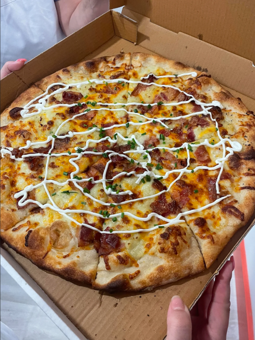 A freshly baked pizza in a cardboard box, topped with cheese, bacon, green onions, and drizzled with mayonnaise.