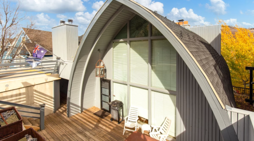 A modern, curved-roof house with large windows, surrounded by trees and a wooden deck with two white chairs.
