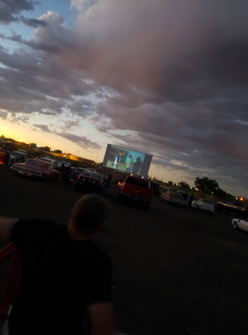 A sunset view of a drive-in movie screen with clouds and parked cars in the foreground.