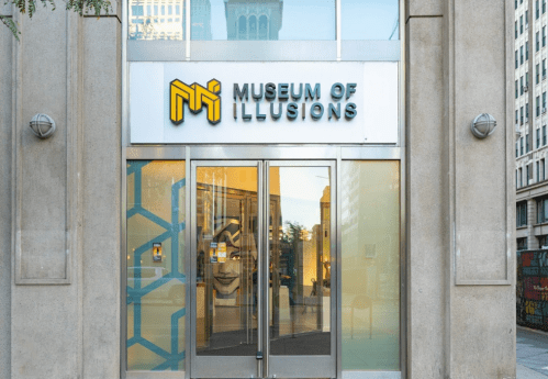 Entrance of the Museum of Illusions, featuring a modern sign and large glass doors.
