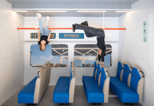 Two people are hanging upside down from a bar in a train-like setting, with blue seats and a scenic backdrop.