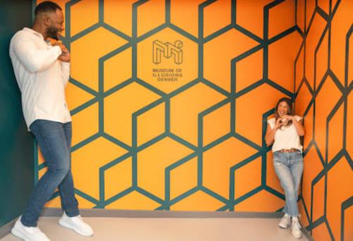 Two people pose playfully in a colorful corner of the Museum of Illusions in Denver, surrounded by geometric patterns.