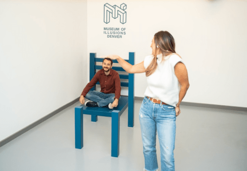A woman playfully gestures towards a man sitting on a large chair at the Museum of Illusions in Denver.