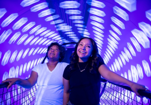 Two girls enjoy a colorful, spinning ride, smiling as vibrant lights swirl around them.