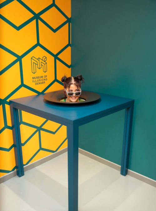 A child with sunglasses peeks out from a circular cutout in a table, set against a colorful wall at the Museum of Illusions.