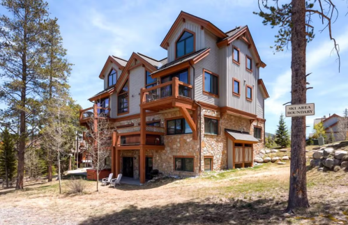 A large, modern mountain house with stone and wood accents, surrounded by trees and a clear blue sky.