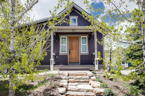 A charming purple house with a wooden door, surrounded by greenery and a stone pathway.