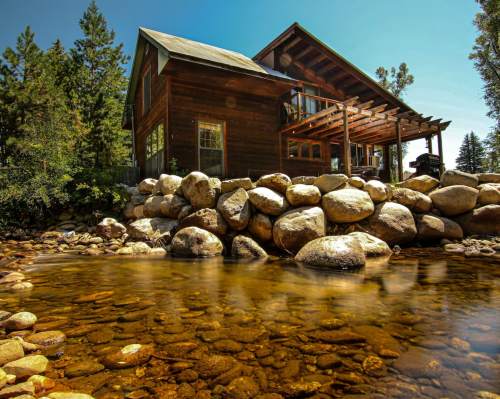 A rustic wooden house by a clear stream, surrounded by trees and large rocks under a bright blue sky.