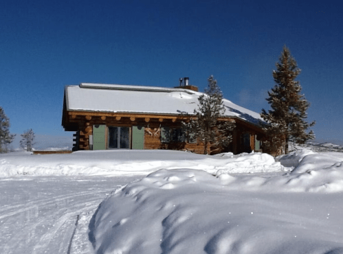 A cozy log cabin surrounded by snow, with clear blue skies and evergreen trees in the background.