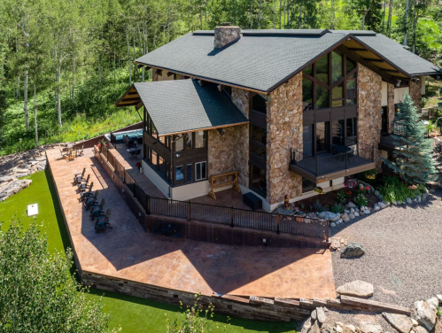 A spacious stone and wood house surrounded by greenery, featuring a large patio and outdoor seating area.