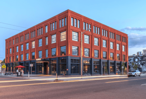 A red brick building with large windows, featuring a café on the ground floor, set against a clear blue sky.