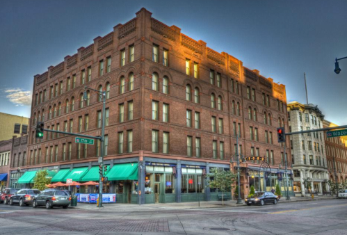 Historic brick building at a street corner, featuring large windows and outdoor seating, with city traffic nearby.