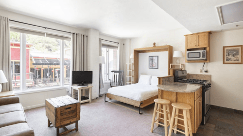 Cozy studio apartment with a bed, kitchenette, TV, and large windows overlooking a street. Warm wood accents throughout.