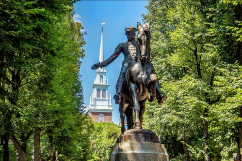 A bronze statue of a mounted figure surrounded by trees, with a church steeple visible in the background.