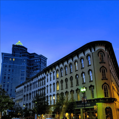 Historic buildings illuminated at dusk, with a clear blue sky and a tall building in the background.