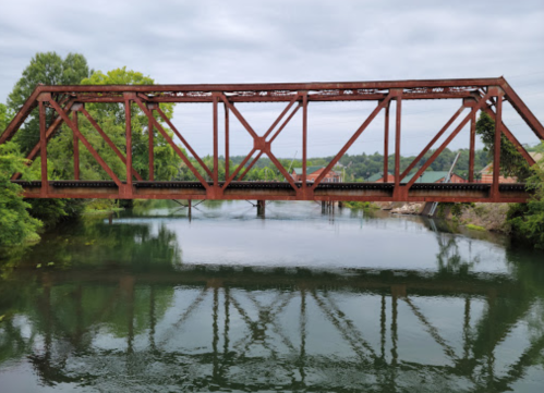 Rusty metal bridge over a calm river, surrounded by greenery and reflecting in the water below.