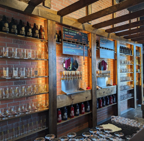 A rustic bar with wooden shelves displaying beer taps, glasses, and a chalkboard menu on a brick wall.
