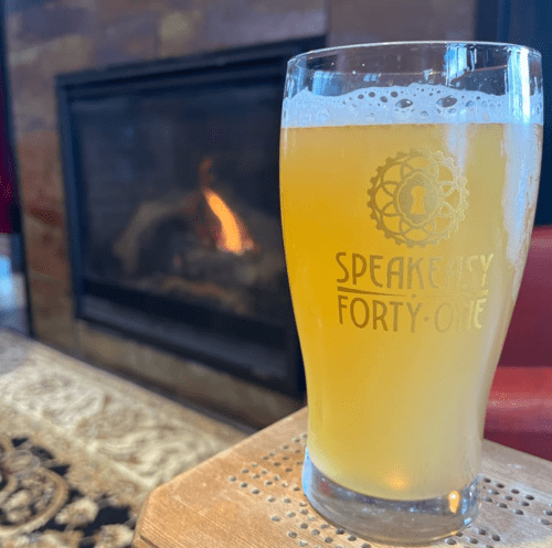 A glass of beer sits on a wooden table in front of a cozy fireplace, with a decorative rug in the background.