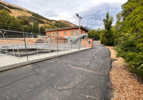 A paved path leads to a fenced facility surrounded by trees and hills under a cloudy sky.