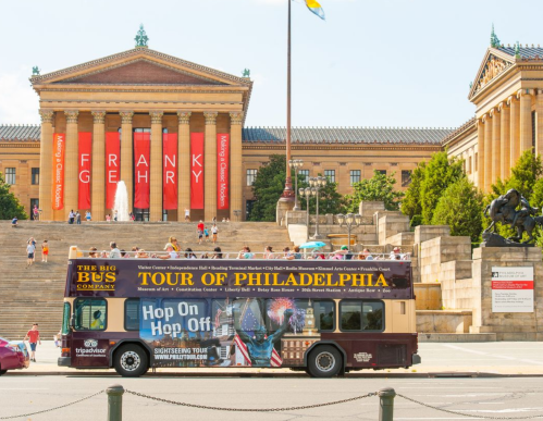 A sightseeing bus in front of the Philadelphia Museum of Art, with steps and greenery in the background.